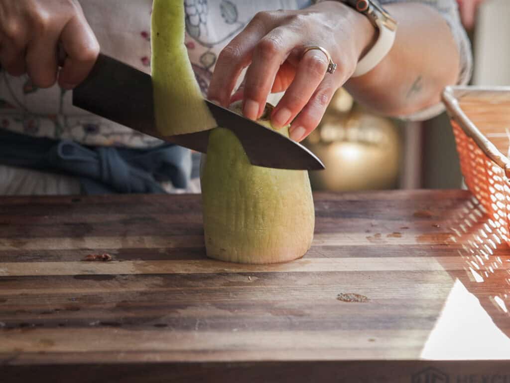 A person slicing the peel of a Korean radish on a wooden cutting board. They are using a large knife and wearing a smartwatch and a ring. The vegetable is upright, partially peeled, with a basket in the background.